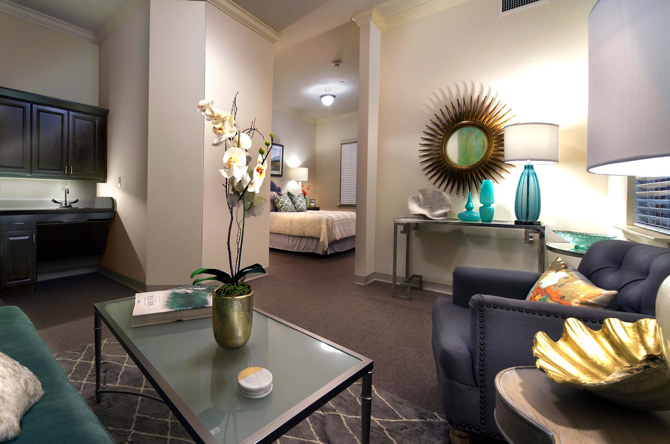 A furnished senior living apartment with a sitting area, kitchenette, and bedroom at The Magnolia at Oxford Commons near Memphis, TN.