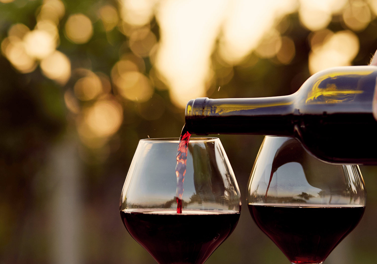 Red wine is poured from a bottle into two stemmed glasses outdoors at sunset, creating a warm, bokeh-filled background.