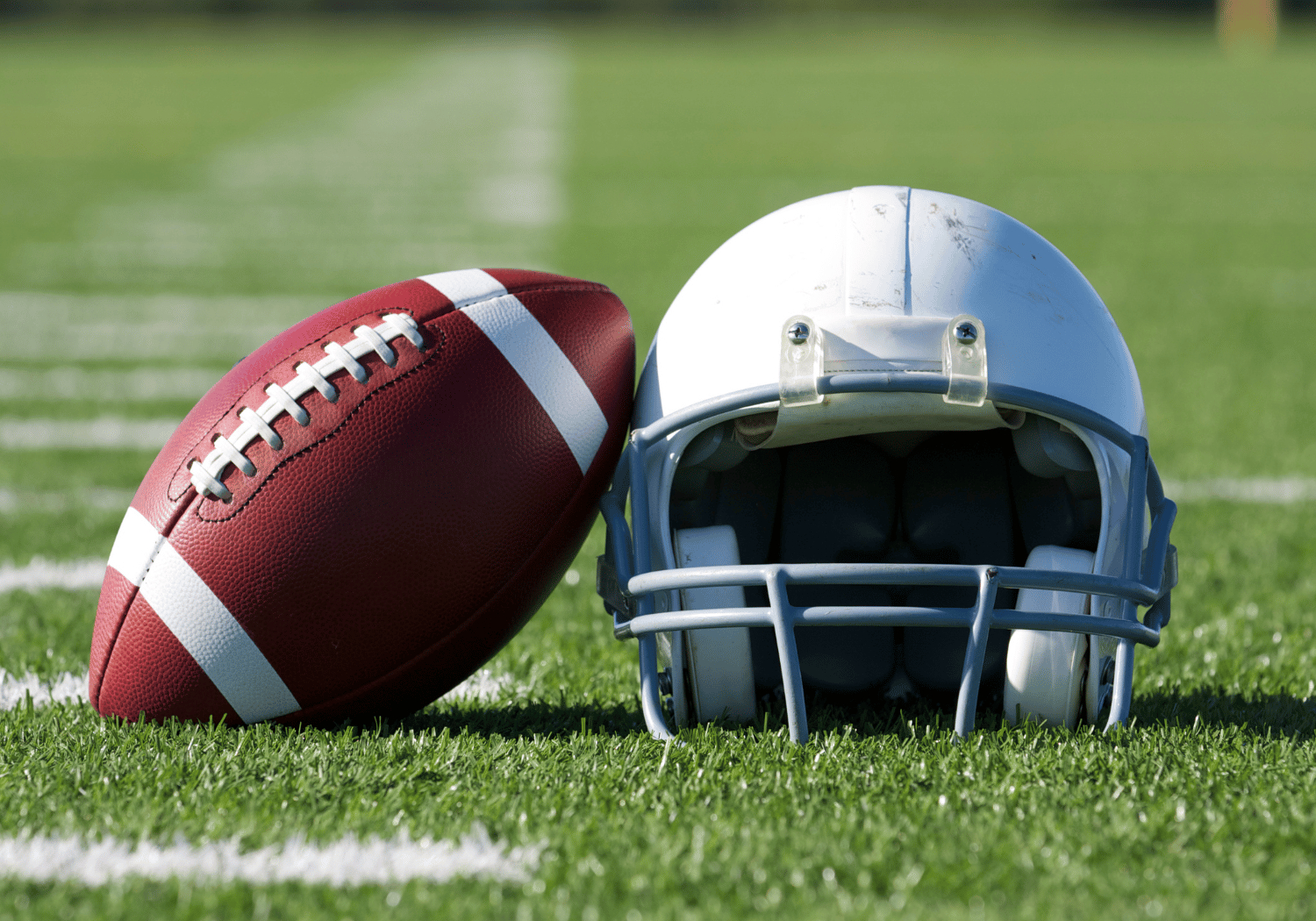 Red football with white stripes beside a white football helmet on a grassy field, ready for play, outdoor sports scene.