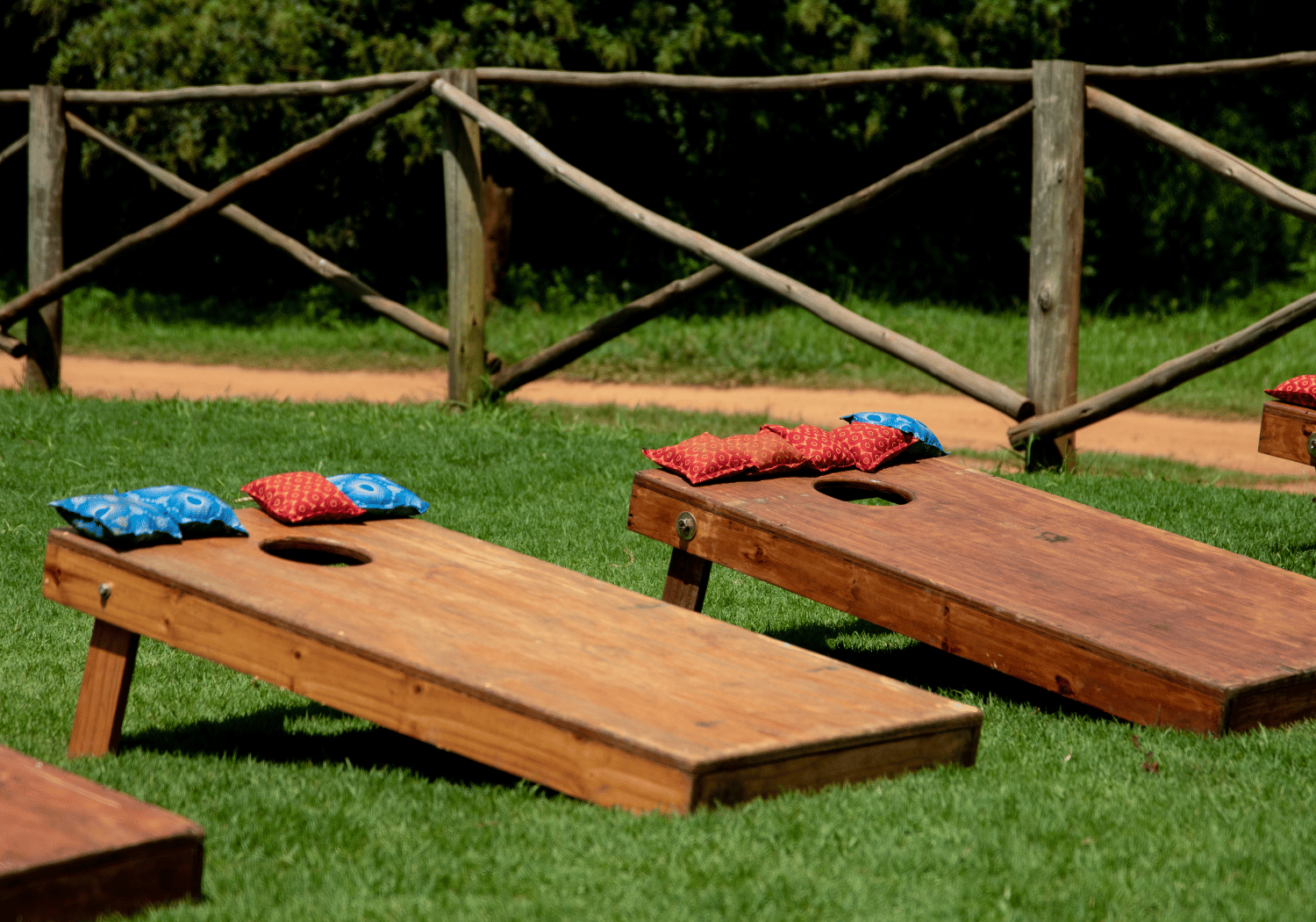 Outdoor cornhole boards on a grassy lawn with colorful bean bags (red and blue) resting on top, wooden fence in the background.
