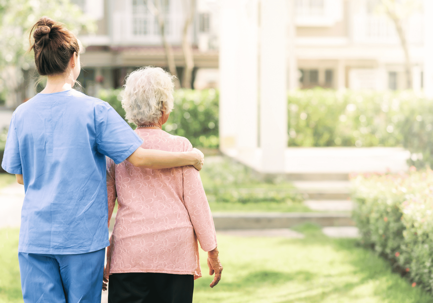 Caregiver in blue scrubs walks with an elderly woman in a pink sweater, arm around her, in a sunny garden.