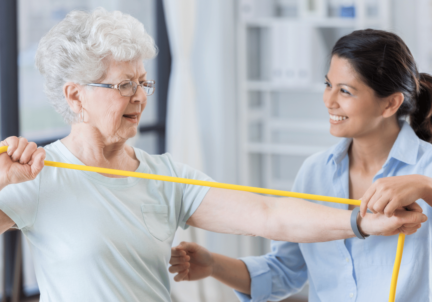 Older woman with white hair doing a shoulder stretch with a yellow resistance band, assisted by a caregiver in a bright room.